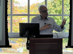 A man stands at a podium in front of large windows while lecturing to conference attendees.