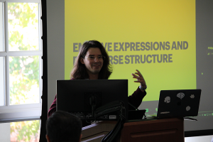 A man with long hair stands behind a podium presenting a PowerPoint slide to conference attendees.