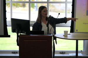 A woman stands at a podium in front of large windows while lecturing to conference attendees.