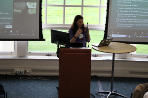 A woman stands at a podium in front of large windows while lecturing to conference attendees.