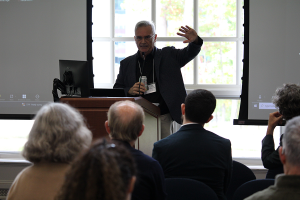 A man stands at a podium in front of large windows while lecturing to conference attendees.