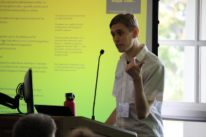 A man stands behind a podium presenting a PowerPoint slide to conference attendees.